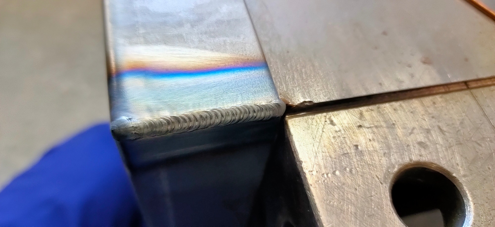 Szczegółowe zbliżenie spoiny TIG na stali nierdzewnej, widoczne charakterystyczne tęczowe przebarwienia termiczne i precyzyjne, drobne ściegi spawalnicze.