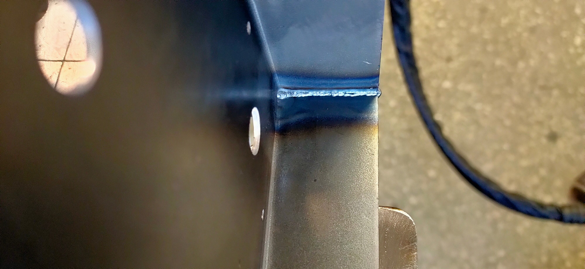 Szczegółowe zbliżenie na spoinę łączącą dwie metalowe płyty pod kątem prostym, widoczne otwory montażowe i ślady po procesie obróbki cieplnej na metalu, w tle fragment przewodu spawalniczego.