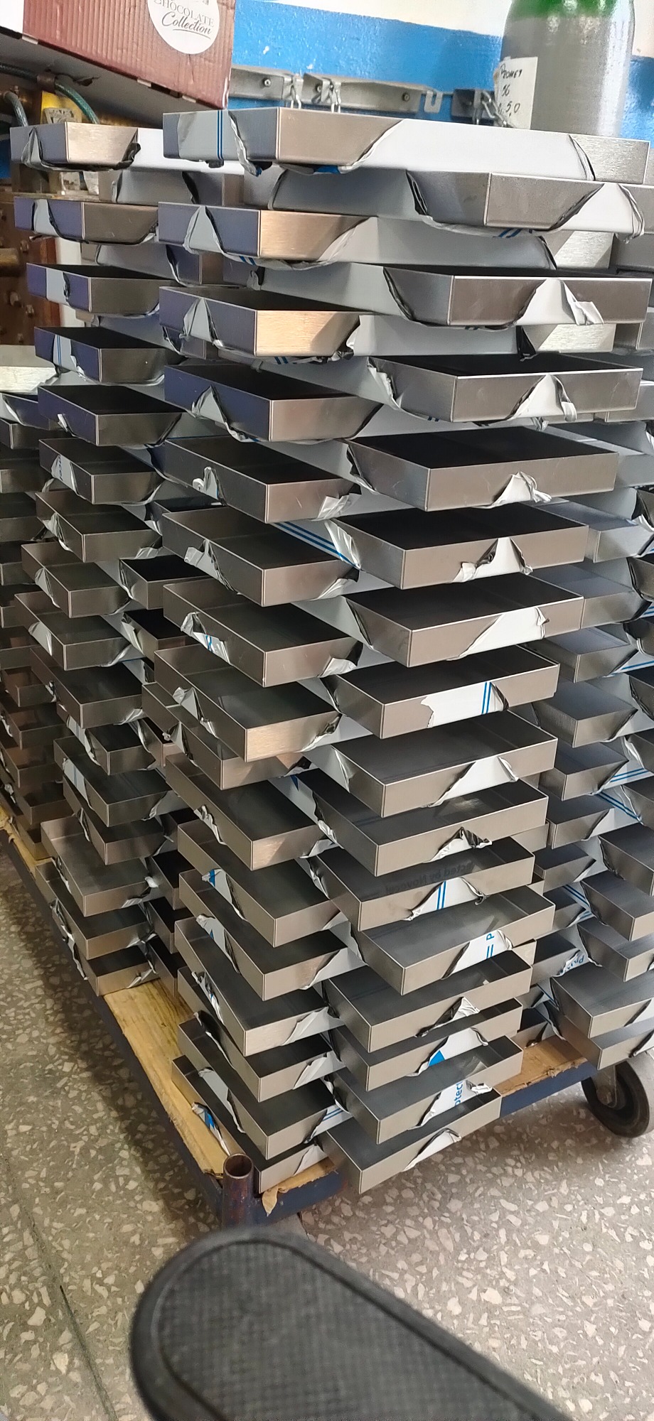 Stos metalowych, prostokątnych pojemników z zabezpieczeniem folią ochronną, ułożonych na palecie transportowej w hali produkcyjnej.