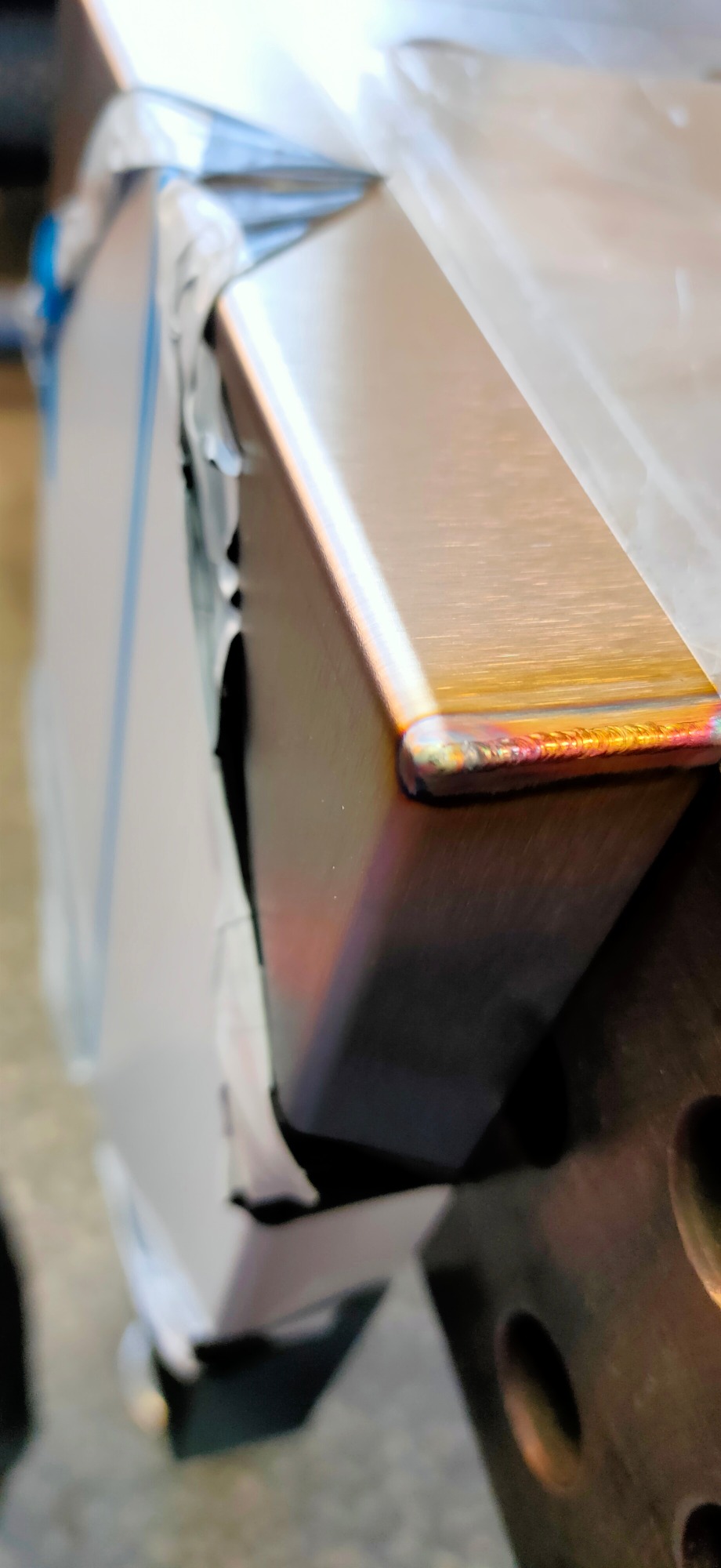 Szlifowana krawędź metalowego profilu zamkniętego, widoczne ślady spawania z charakterystycznym zabarwieniem termicznym, częściowo zabezpieczonego folią ochronną.