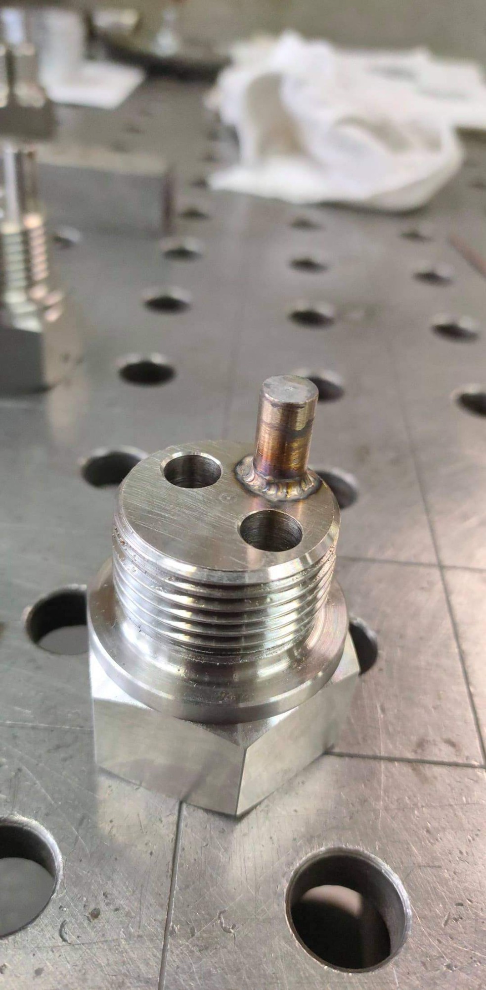 Metalowy element z trzema otworami i przyspawanym pionowym prętem, leżący na perforowanym stole spawalniczym. Widoczne ślady po spawaniu na połączeniu pręta i elementu.