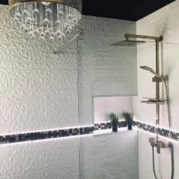 Nowoczesna kabina prysznicowa z białymi trójwymiarowymi płytkami, złotą armaturą, podświetlaną wnęką z dekoracyjnymi roślinami i kryształowym żyrandolem.