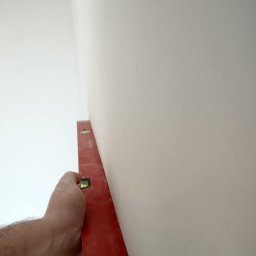 Platynowa Budowa Adrian Kania - Czerwona poziomica przyłożona do świeżo pomalowanej, jasnej ściany w rogu pomieszczenia, sprawdzanie pionu