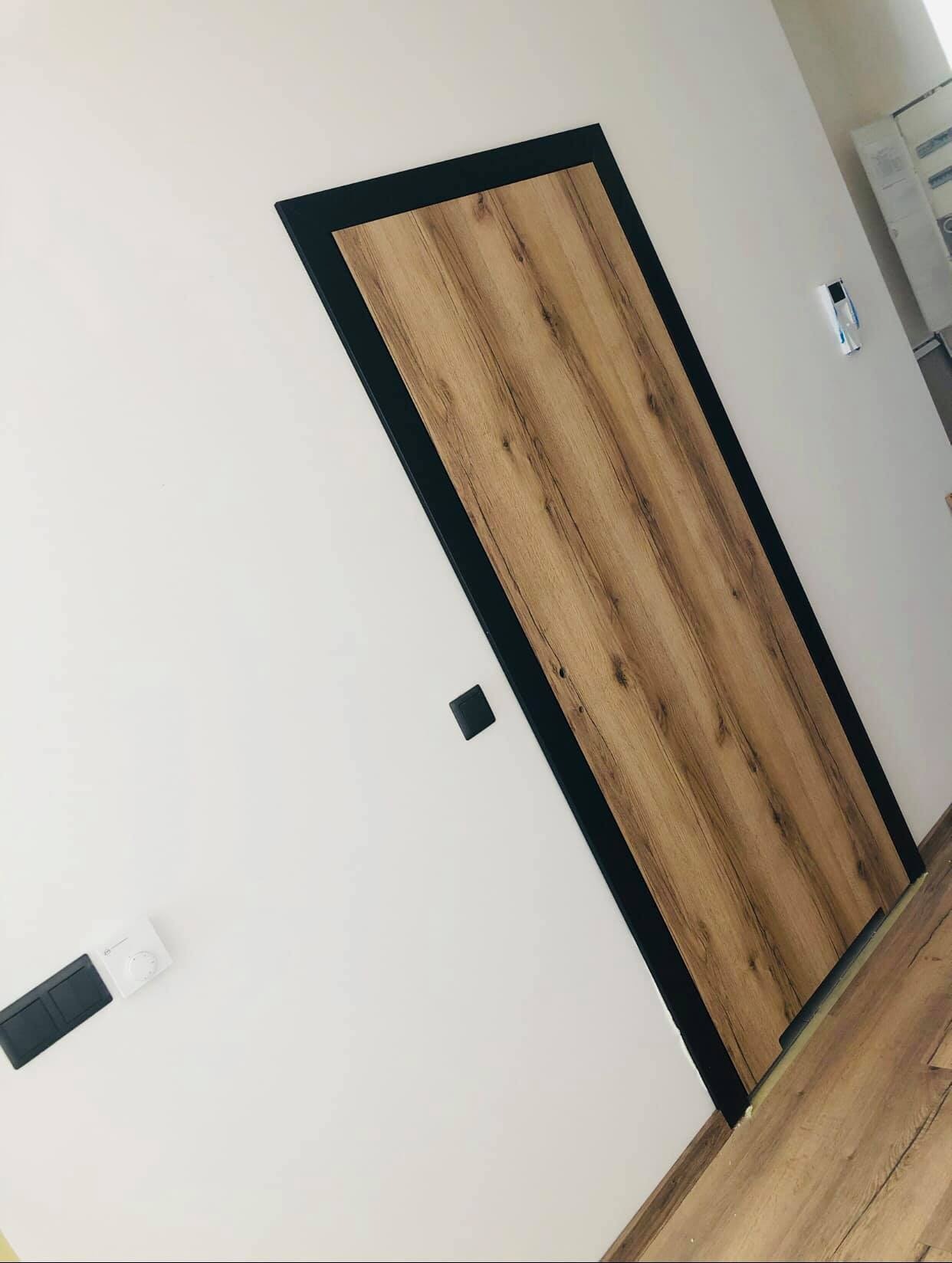 Zamontowane drzwi wewnętrzne o wzorze drewna i czarnej ramie, widoczne na białej ścianie z włącznikami i regulatorem temperatury.