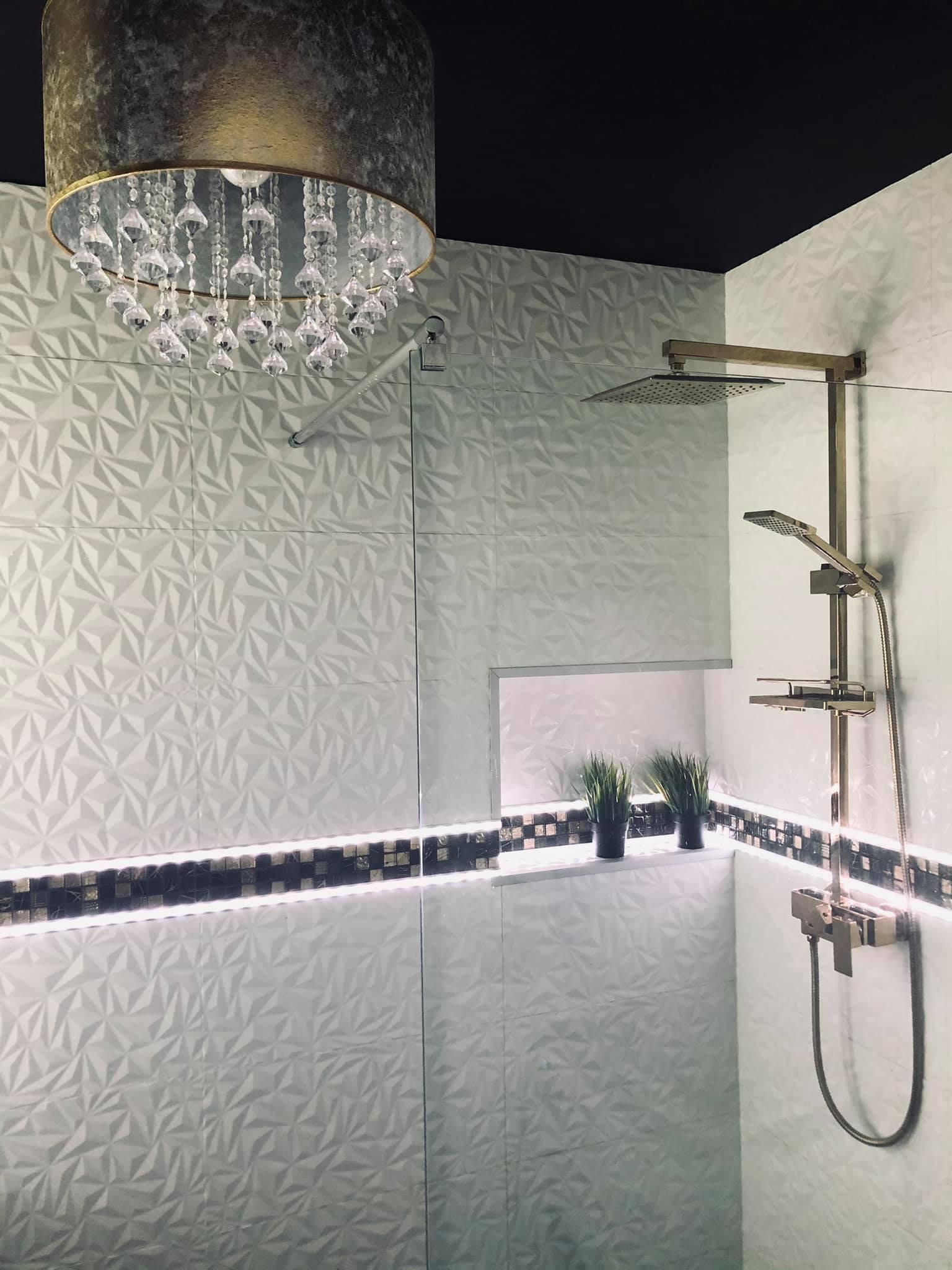 Nowoczesna kabina prysznicowa z białymi trójwymiarowymi płytkami, złotą armaturą, podświetlaną wnęką z dekoracyjnymi roślinami i kryształowym żyrandolem.