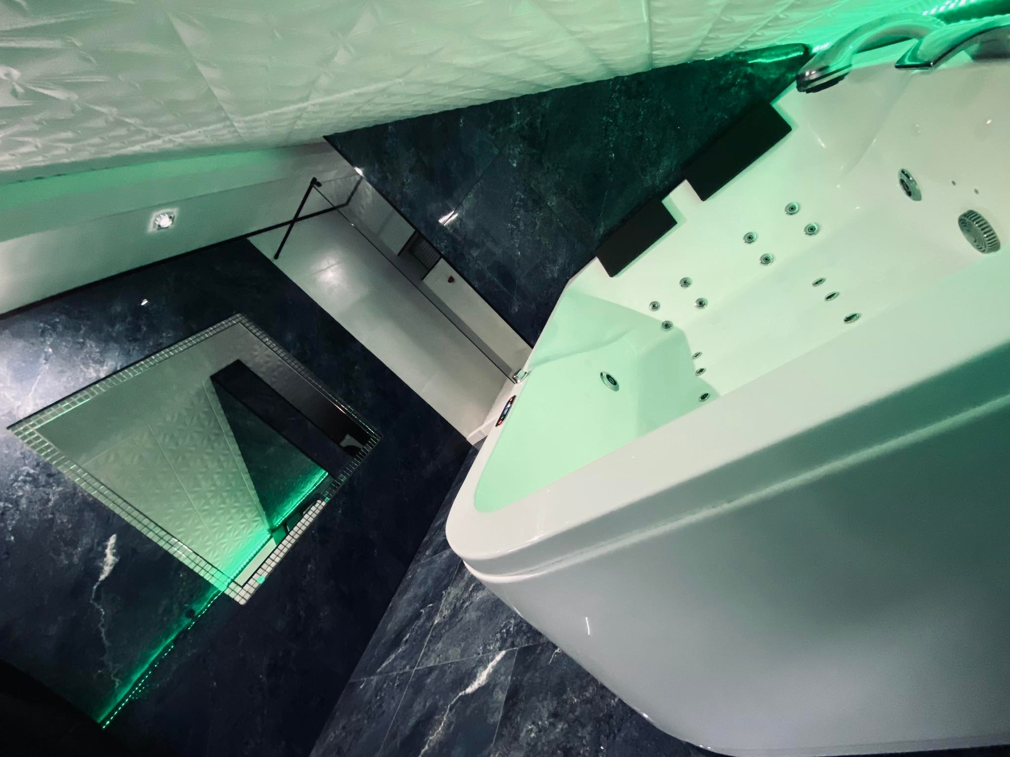 Nowoczesna łazienka z marmurowymi płytkami, podświetlanym lustrem i białą wanną z hydromasażem, oświetlona zielonymi lampami LED.