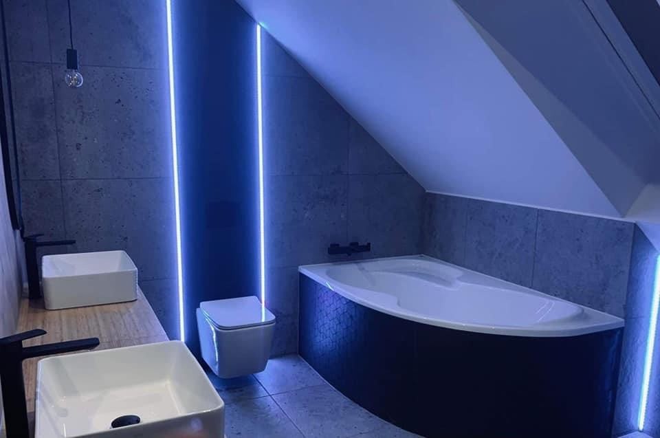 Nowoczesna łazienka na poddaszu z umywalkami nablatowymi, podwieszaną toaletą i narożną wanną z mozaiką, oświetlona pionowymi pasami LED.