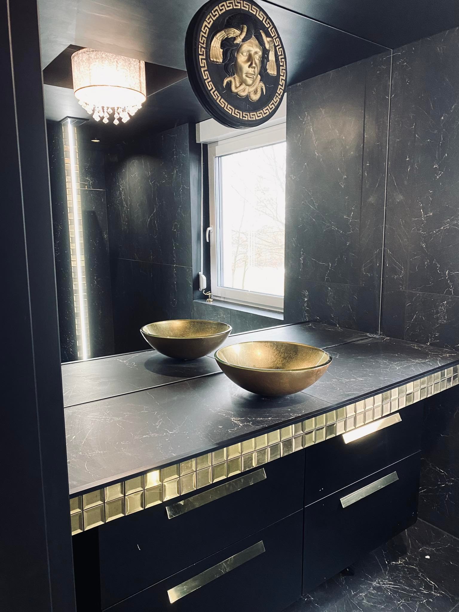 Elegancka łazienka w ciemnej tonacji z marmurowymi płytkami, złotymi umywalkami w kształcie mis, dekoracyjnym oświetleniem i ozdobnym medalionem na ścianie.