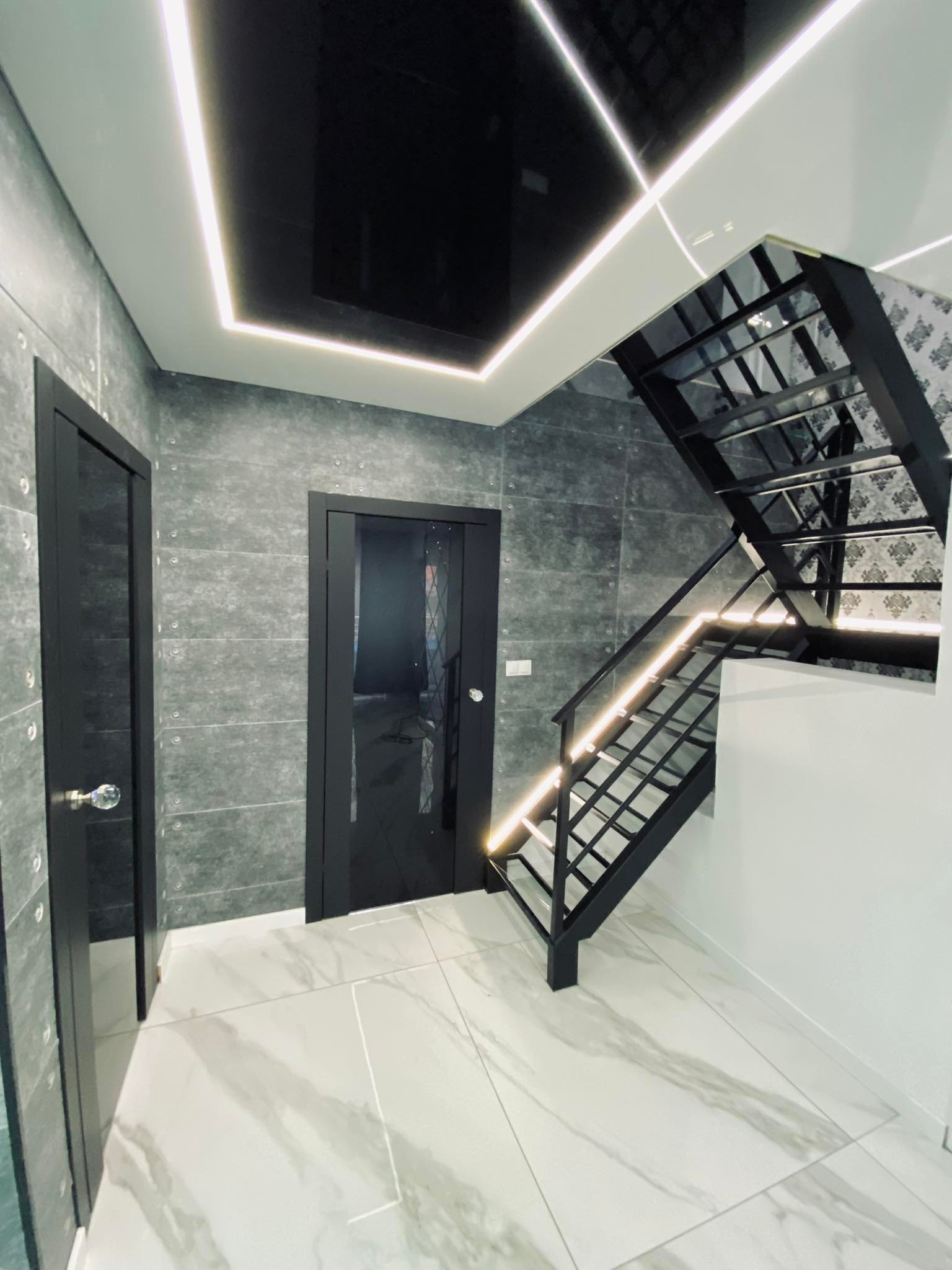 Nowoczesne wnętrze: czarne schody z podświetleniem LED, szare płytki imitujące beton na ścianach, biała marmurowa podłoga i czarne drzwi z kryształową gałką.