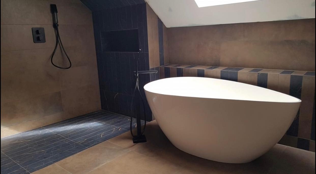 Nowoczesna łazienka na poddaszu z wolnostojącą białą wanną, czarną armaturą i płytkami w odcieniach beżu i granatu.