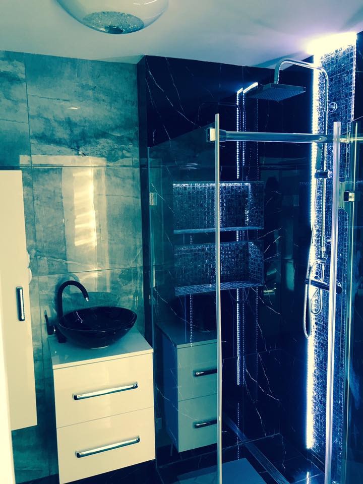 Nowoczesna łazienka z czarnymi marmurowymi płytkami, szklaną kabiną prysznicową z oświetleniem LED, białą szafką pod umywalką z czarną misą i designerską lampą sufitową.