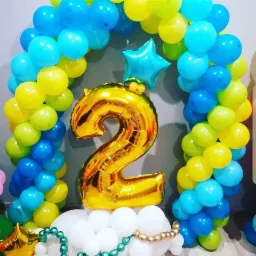 Dekoracja balonowa na drugie urodziny: złoty balon w kształcie cyfry 2, łuk z niebieskich i żółtych balonów, oraz biała podstawa z balonów, całość na tle szarej ściany.