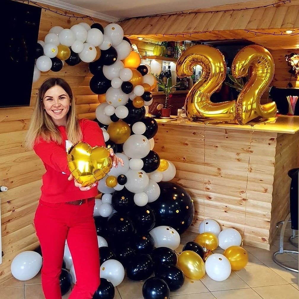 Uśmiechnięta kobieta w czerwonym stroju trzyma złoty balon w kształcie serca na tle dekoracji z białych, czarnych i złotych balonów oraz złotych cyfr '22' na drewnianym barze.