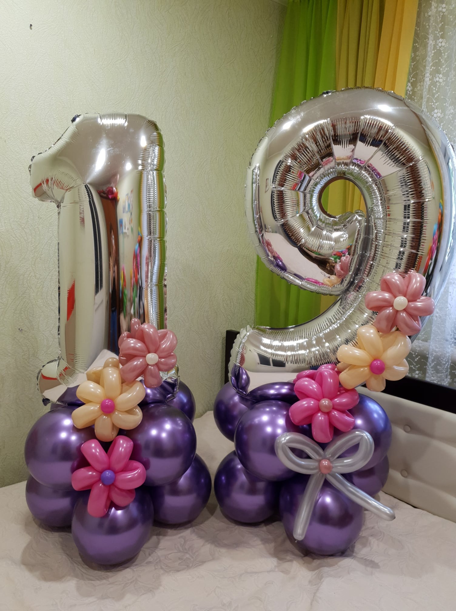 Dwa srebrne balony foliowe w kształcie cyfr '1' i '9', tworzące liczbę 19, udekorowane fioletowymi balonami i kwiatami z balonów w kolorach różowym i brzoskwiniowym, ustawione na jasnym tle.