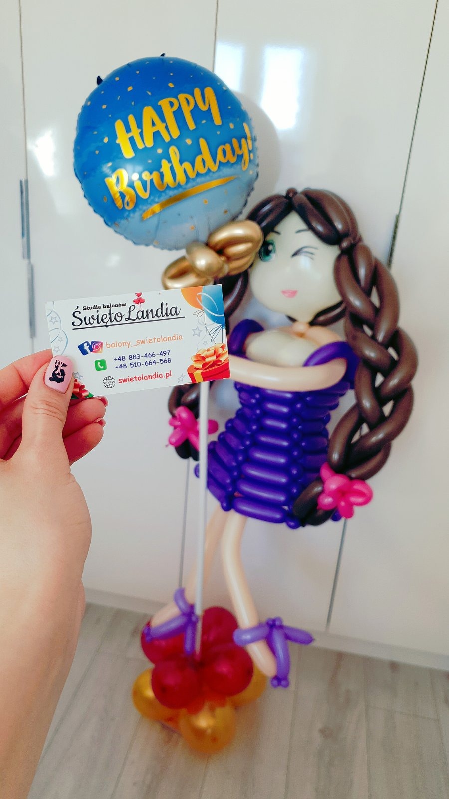 Figurka dziewczyny z balonów w fioletowej sukience, z niebieskim balonem 'Happy Birthday' nad głową, obok dłoń trzymająca wizytówkę Studia Balonów Święto Landia.