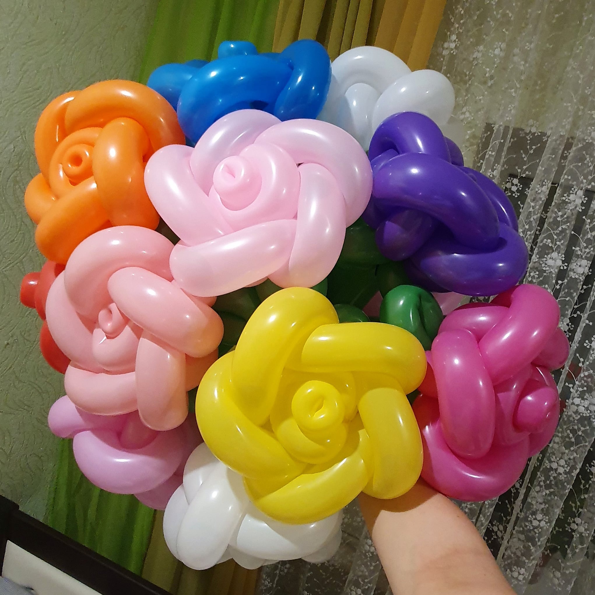 Bukiet kolorowych kwiatów uformowanych z długich balonów, trzymany w dłoni na tle zielonej ściany i firanki z kwiatowym wzorem.