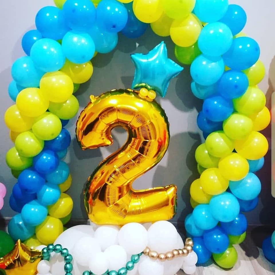Dekoracja balonowa na drugie urodziny: złoty balon w kształcie cyfry 2, łuk z niebieskich i żółtych balonów, oraz biała podstawa z balonów, całość na tle szarej ściany.