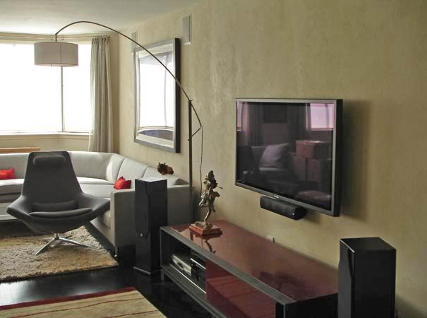 Salon z telewizorem na ścianie, szarą sofą narożną, fotelem obrotowym w stylu retro, lampą łukową i ścianą pokrytą dekoracyjnym tynkiem w odcieniach beżu.