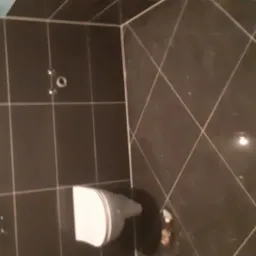 Ciemne, błyszczące płytki w łazience z widoczną muszlą klozetową i instalacją pod umywalkę, częściowo ukończone wykończenie.