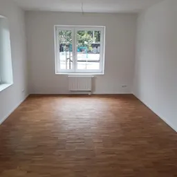 Pusty pokój po remoncie z parkietem w jodełkę, świeżo pomalowane białe ściany, widoczne kable zwisające z sufitu, okno z widokiem na zieleń.