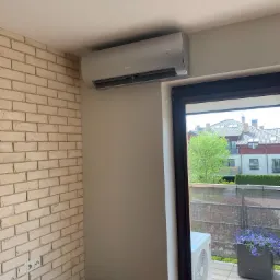 Nowoczesny klimatyzator ścienny marki Gree zamontowany w jasnym pomieszczeniu z ceglaną ścianą i widokiem na balkon z kwiatami.