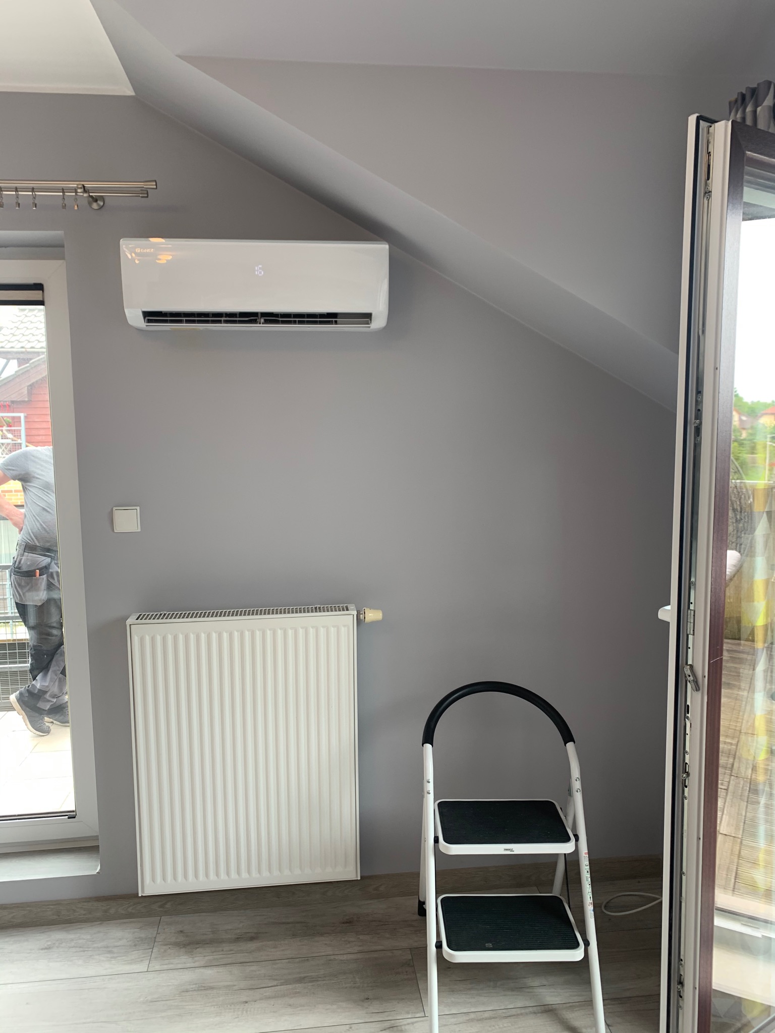 Biały klimatyzator ścienny zamontowany w szarym pokoju na poddaszu, drabina dwustopniowa stoi obok otwartego okna balkonowego, widoczny fragment instalatora.