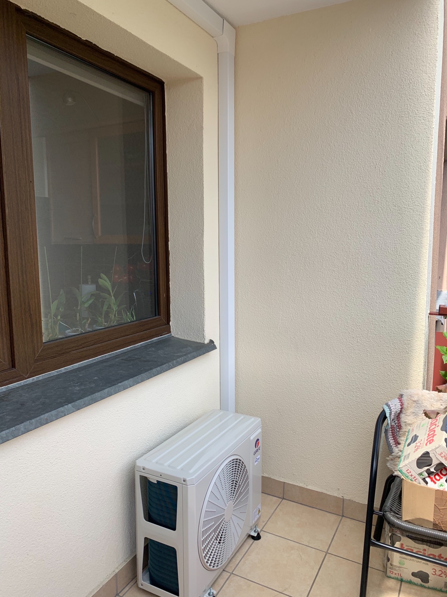 Zewnętrzna jednostka klimatyzatora zamontowana na balkonie przy oknie z brązową ramą, obok widoczny fragment krzesła z kartonami mleka, ściana w kolorze kremowym.