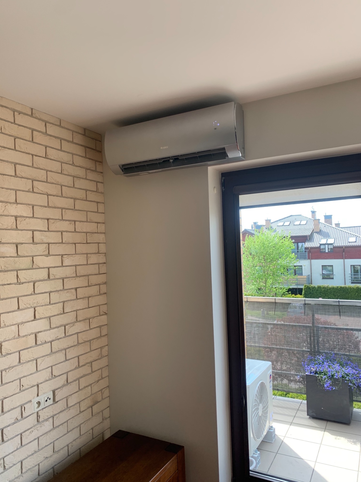 Nowoczesny klimatyzator ścienny marki Gree zamontowany w jasnym pomieszczeniu z ceglaną ścianą i widokiem na balkon z kwiatami.