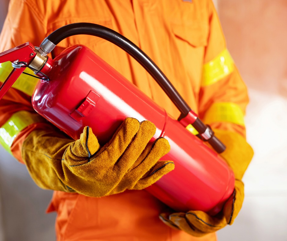 Osoba w pomarańczowym kombinezonie ochronnym i rękawicach trzyma czerwony gaśnicę proszkową.