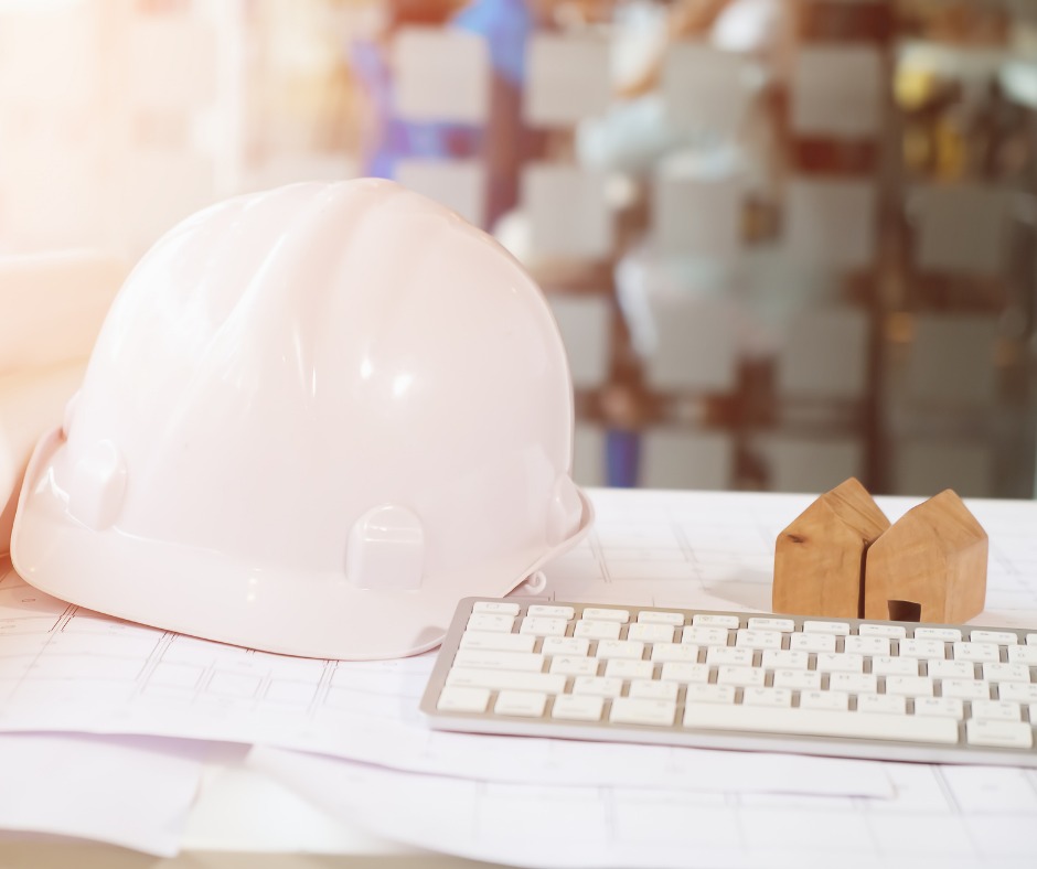 Biały kask ochronny, klawiatura i drewniane modele domów na stole z rozłożonymi planami budowlanymi, w tle rozmyta szklana ściana.