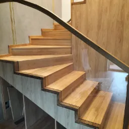 Nowoczesne schody drewniane z ciemną poręczą i szklanymi panelami bocznymi, zintegrowane z zabudową pod schodami w odcieniach drewna.