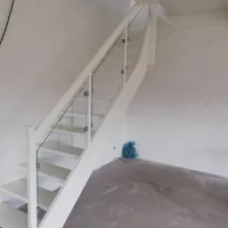 Białe schody z przezroczystą szklaną balustradą i metalowymi mocowaniami, widok z dołu, na podłodze niebieski worek na śmieci.