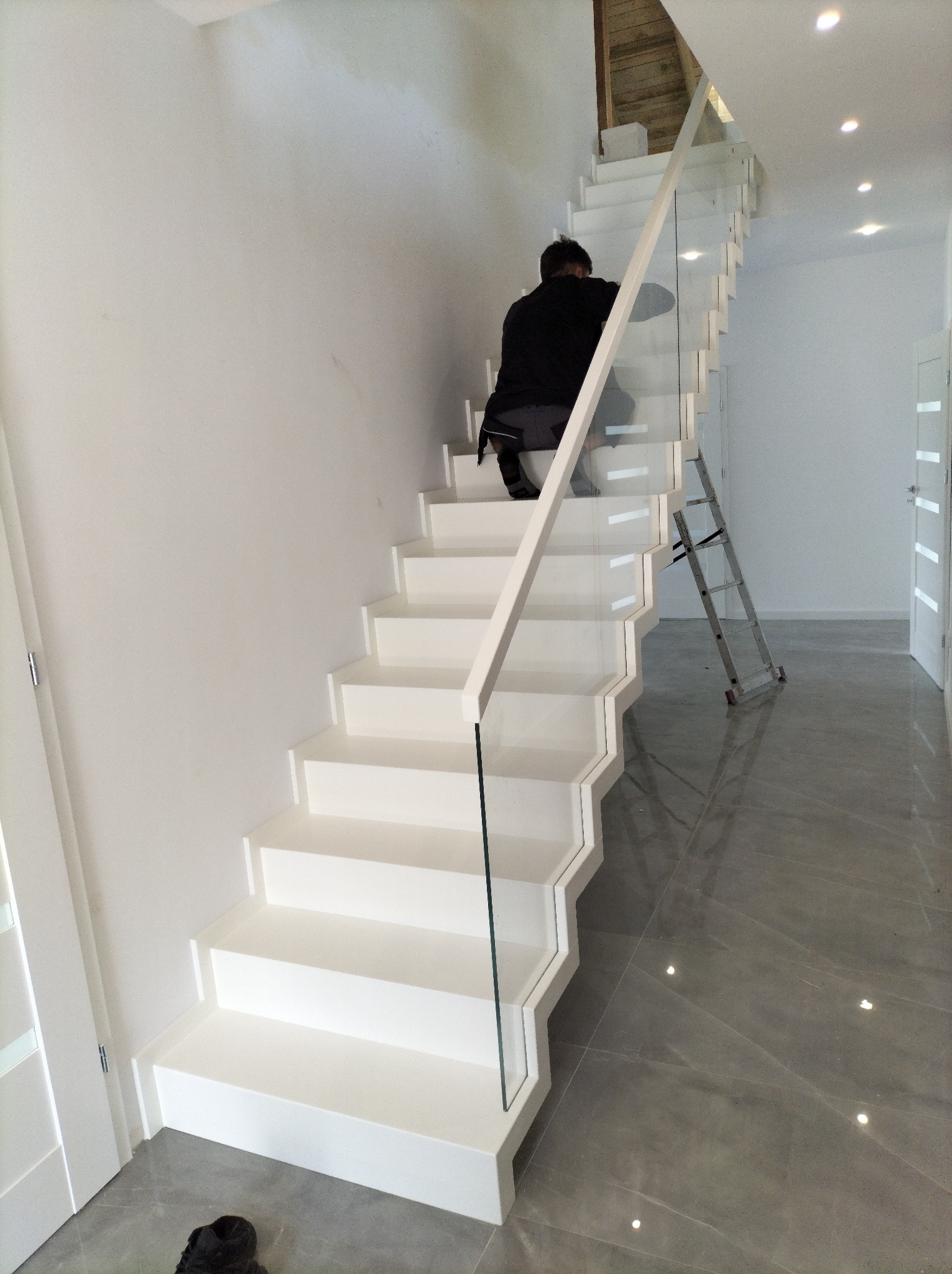Białe schody z szklaną balustradą w nowoczesnym wnętrzu. Widoczna osoba na schodach i drabina oparta o balustradę. Minimalistyczny design i jasna kolorystyka.