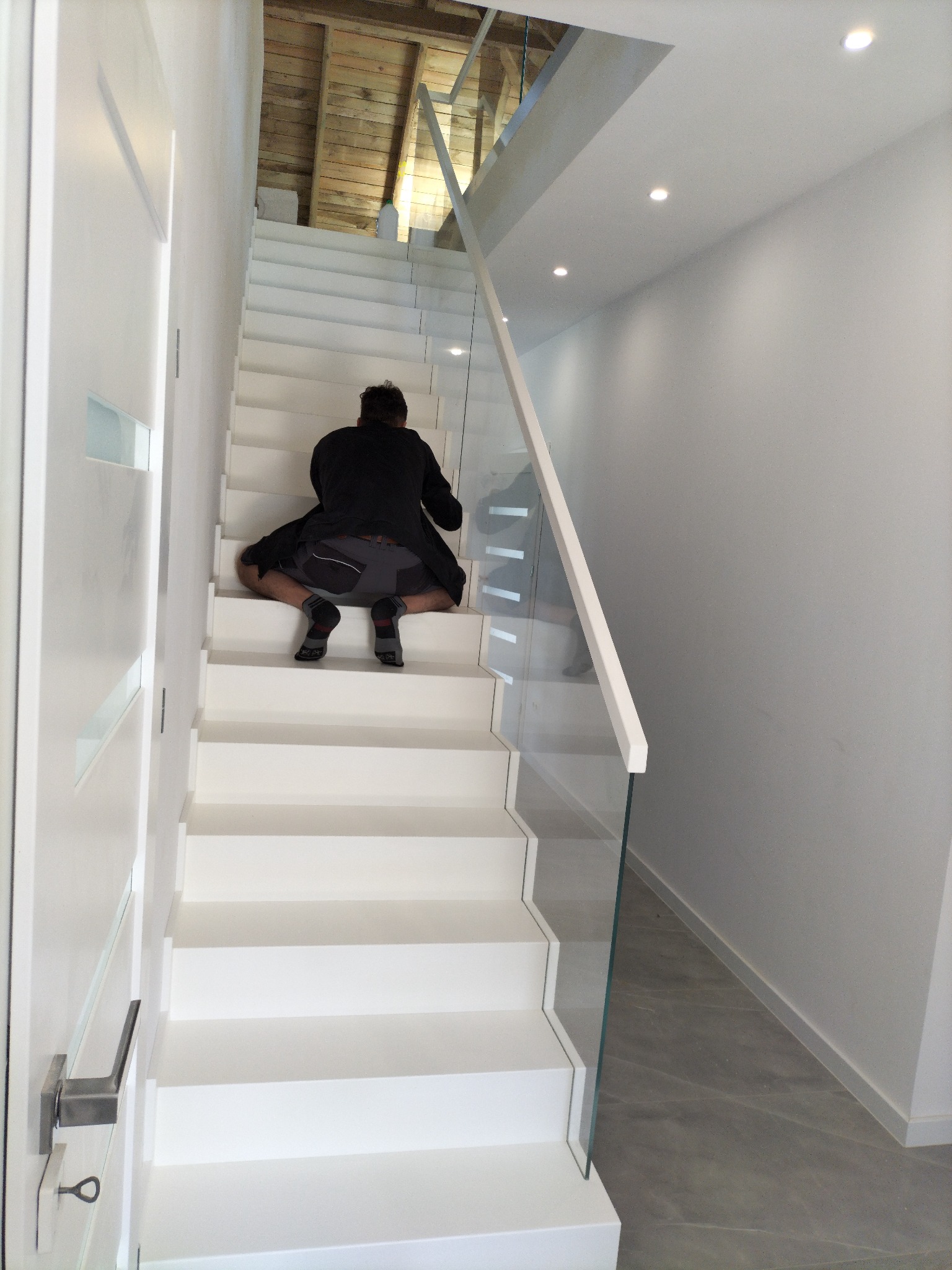 Białe schody z szklaną balustradą w nowoczesnym wnętrzu. Mężczyzna w czarnej bluzie siedzi na schodach tyłem do kamery. Widoczne drewniane wykończenie sufitu.