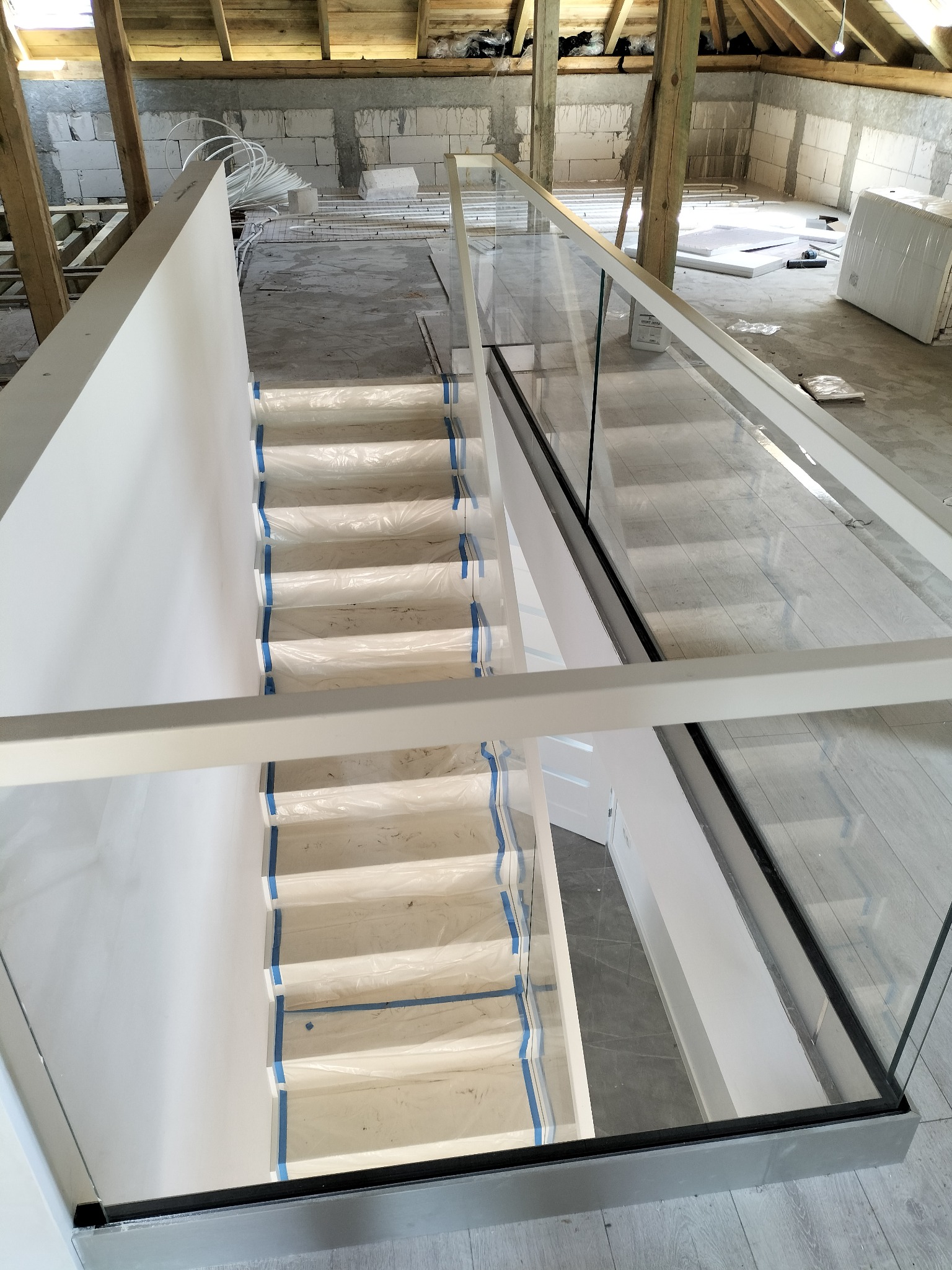 Białe schody z zabezpieczonymi stopniami i szklaną balustradą w surowym wnętrzu poddasza, widoczne elementy konstrukcyjne dachu i instalacje.