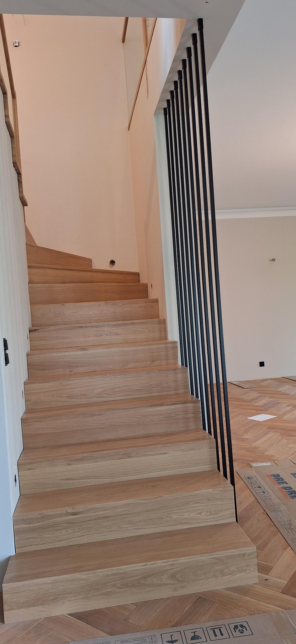 Jasne schody drewniane z czarną, pionową balustradą w nowoczesnym wnętrzu. Parkiet ułożony w jodełkę. Widok z dołu, schody prowadzą na górę.