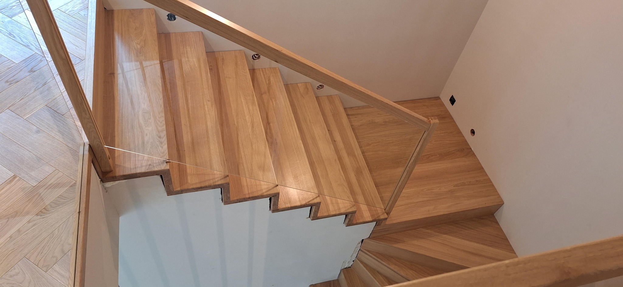 Kątowe ujęcie nowoczesnych schodów z jasnego drewna, z przezroczystą balustradą, oświetleniem punktowym i parkietem w jodełkę. Minimalistyczny design.