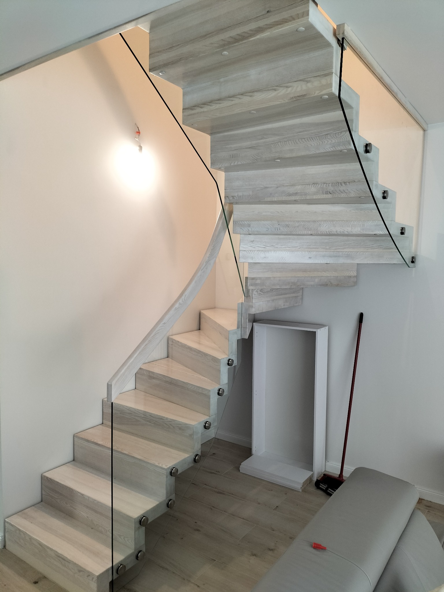 Białe, spiralne schody drewniane z balustradą szklaną, mocowaną punktowo. Widoczne jasne drewno i minimalistyczny design. Ujęcie z dołu, lekko z ukosa.