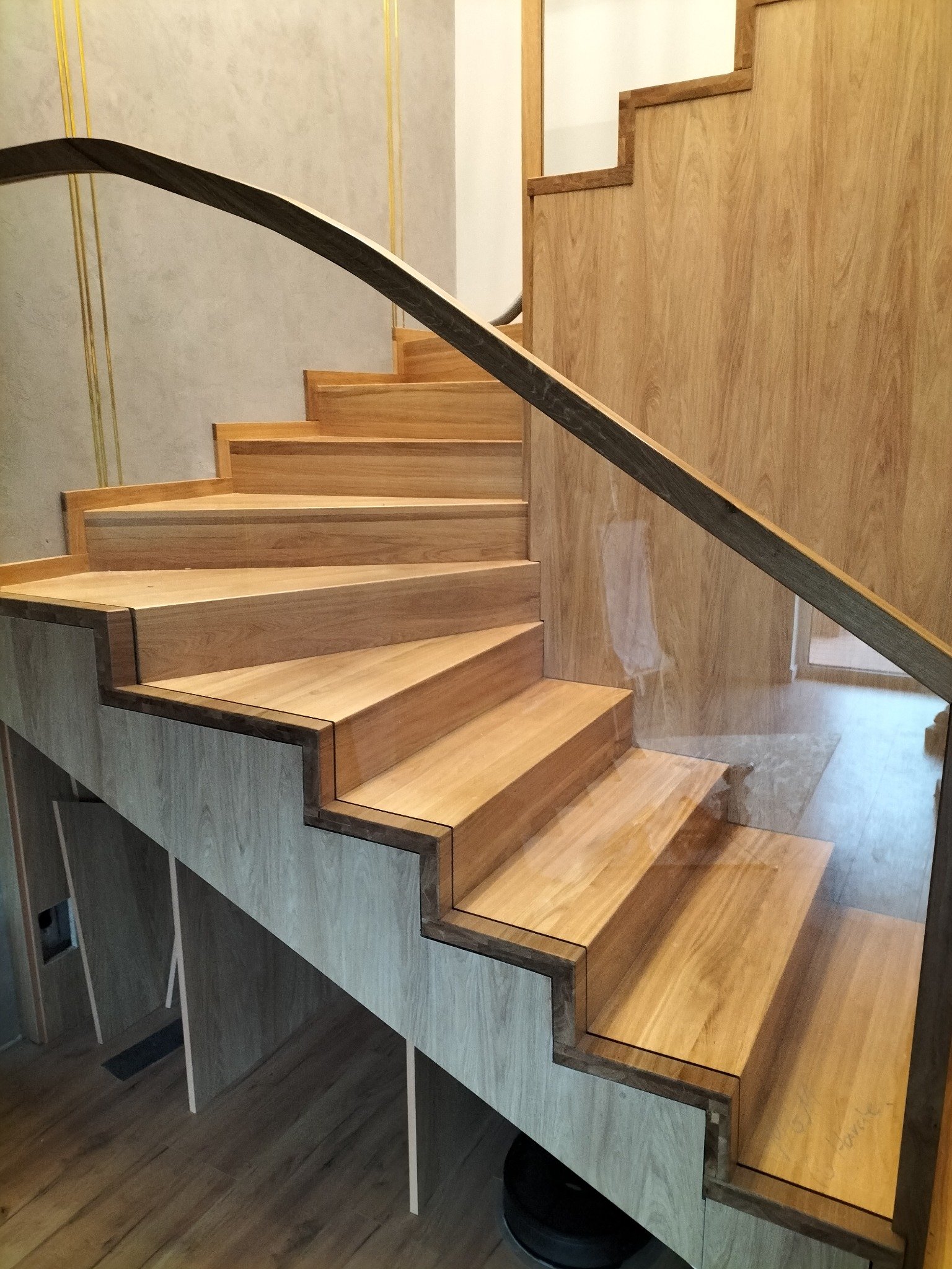 Nowoczesne schody drewniane z ciemną poręczą i szklanymi panelami bocznymi, zintegrowane z zabudową pod schodami w odcieniach drewna.