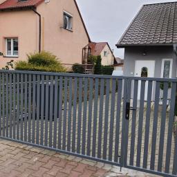 Smart Metal Construction - Nowoczesna, szara brama posesyjna z pionowych profili, widok z zewnątrz na kostkę brukową i zabudowę jednorodzinną w tle.