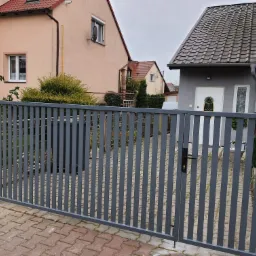 Nowoczesna, szara brama posesyjna z pionowych profili, widok z zewnątrz na kostkę brukową i zabudowę jednorodzinną w tle.