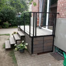 Zewnętrzny taras z drewnianą podłogą i czarną metalową balustradą, prowadzący do drzwi balkonowych w budynku z cegły, z doniczkami z kwiatami na schodach.