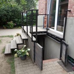 Smart Metal Construction - Zewnętrzne schody z metalową ramą i drewnianymi stopniami prowadzące do piwnicy, z balustradą i roślinami w donicach, widoczne ceglana ściana budynku i fragment ogrodu.