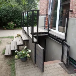 Zewnętrzne schody z metalową ramą i drewnianymi stopniami prowadzące do piwnicy, z balustradą i roślinami w donicach, widoczne ceglana ściana budynku i fragment ogrodu.