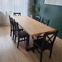 Stół z jasnego drewna z czarnymi krzesłami w pokoju z zieloną ścianą i zasłonami w tle.