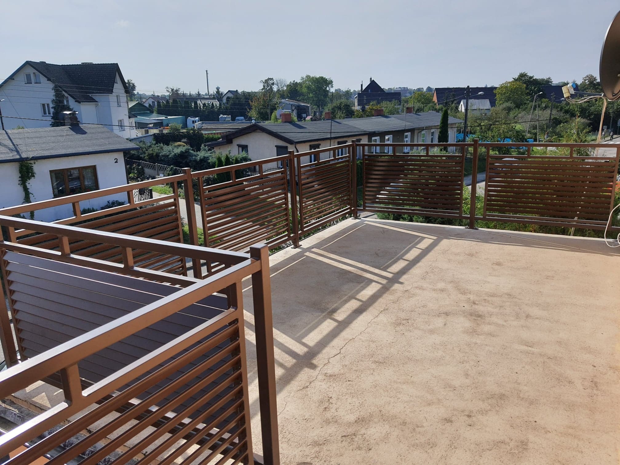 Nowoczesna balustrada balkonowa z poziomych, brązowych profili, widok z góry na tle zabudowy jednorodzinnej i błękitnego nieba.