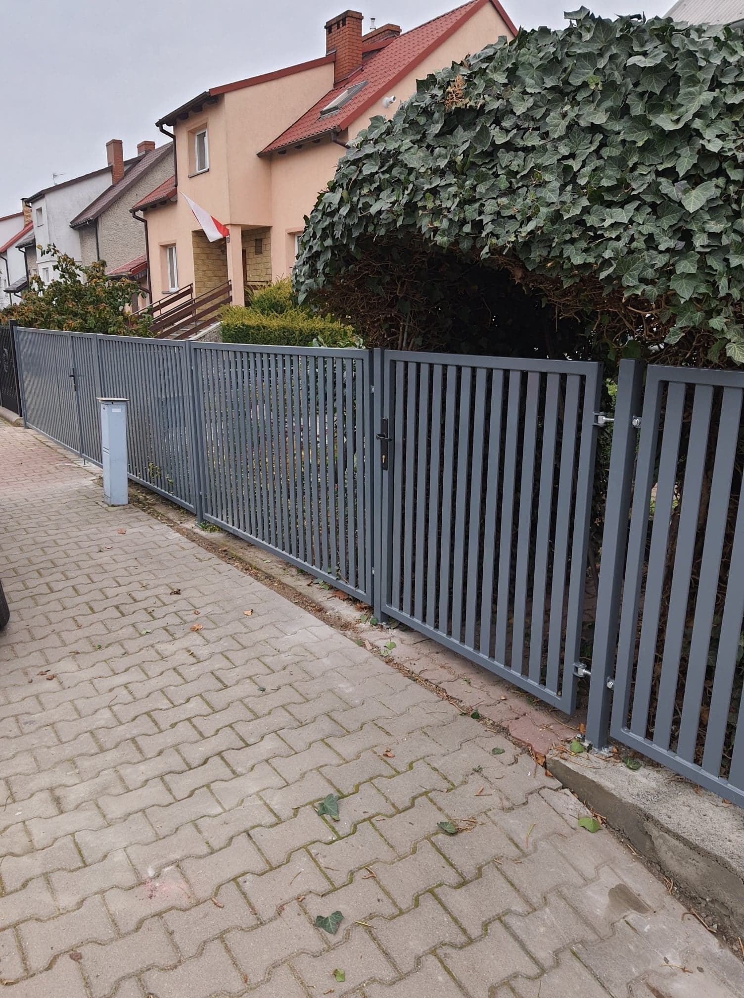 Nowoczesne, szare ogrodzenie z pionowych profili, z bramką z czarnym zamkiem, oddzielające posesję od chodnika z kostki brukowej. W tle dom z polską flagą.