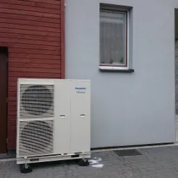 Zewnętrzna jednostka klimatyzacyjna Panasonic Inverter umieszczona na czarnych podkładkach antywibracyjnych, stoi na szarej kostce brukowej przy ścianie budynku z brązowymi drewnianymi panelami...