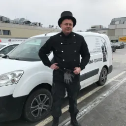 Mężczyzna w stroju roboczym z cylindrem i rękawicami, stojący przed białym samochodem firmowym z numerem telefonu i logotypem, na parkingu przed budynkiem.