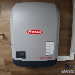 Szary inwerter Fronius Symo zamontowany na ścianie z drewnopodobną okładziną, obok skrzynka elektryczna w białej obudowie. Widoczny ekran z wartością 7750 oraz naklejka informacyjna o monitoringu...